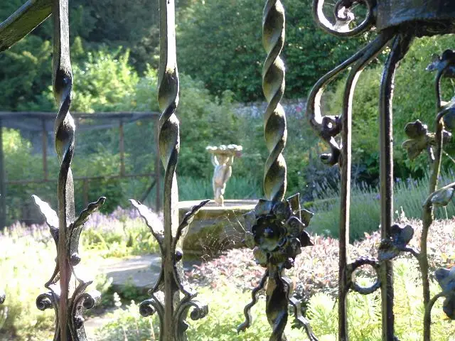 Seen through the bars of a ornate wrought iron gate, a fountain in the walled garden is visible, in front of a fruitcage and shrubs